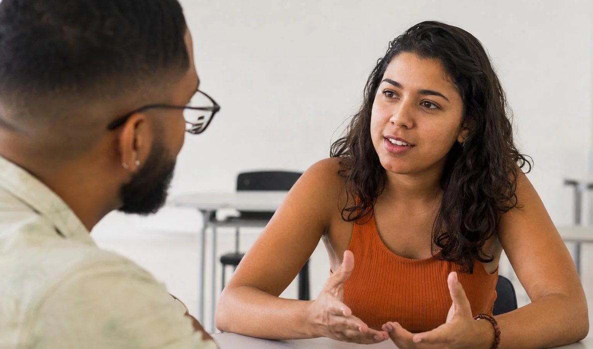 Três participantes conversam ao redor de uma mesa comprida numa sala clara de janelas altas